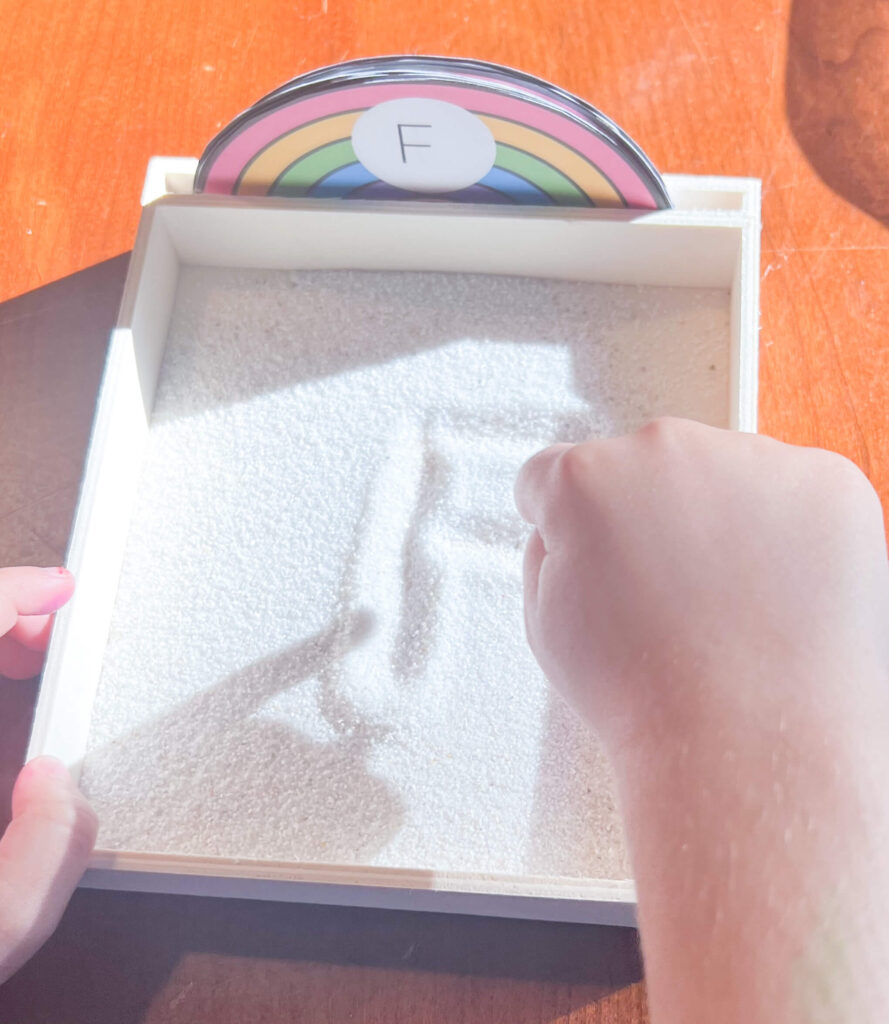 Image shows a sand tray with the letter "F" rainbow card. A hand is above the tray writing the letter F in the sand.