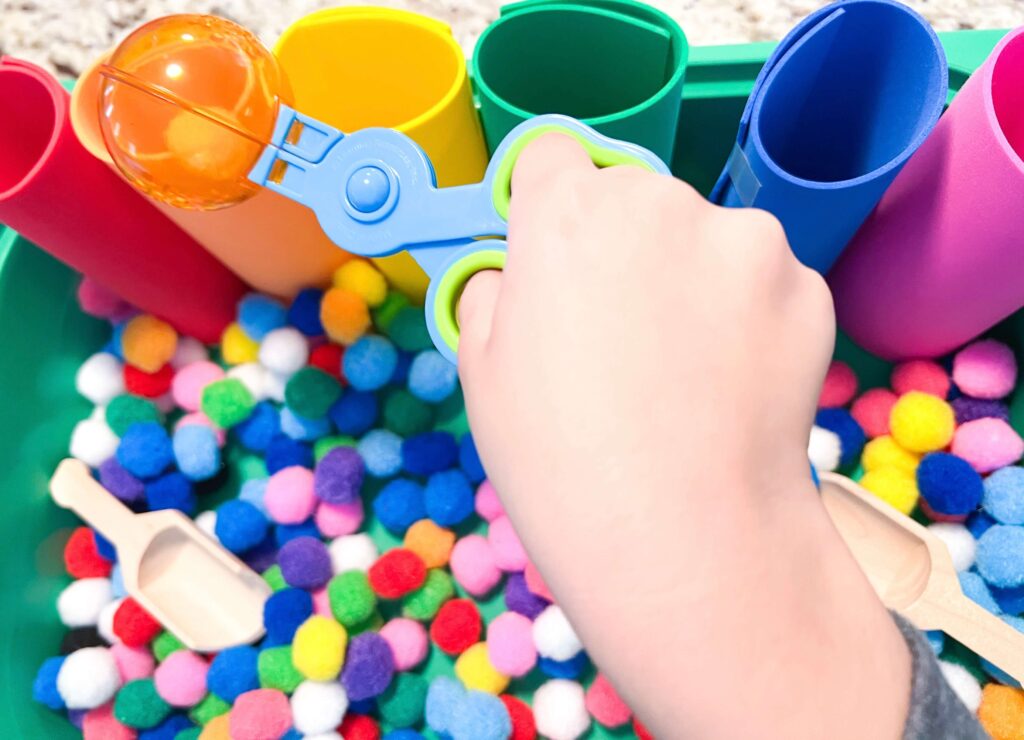 Image shows a sensory bin filled with pom poms and a child's hand is holding a scoop with pom poms inside above the bin.